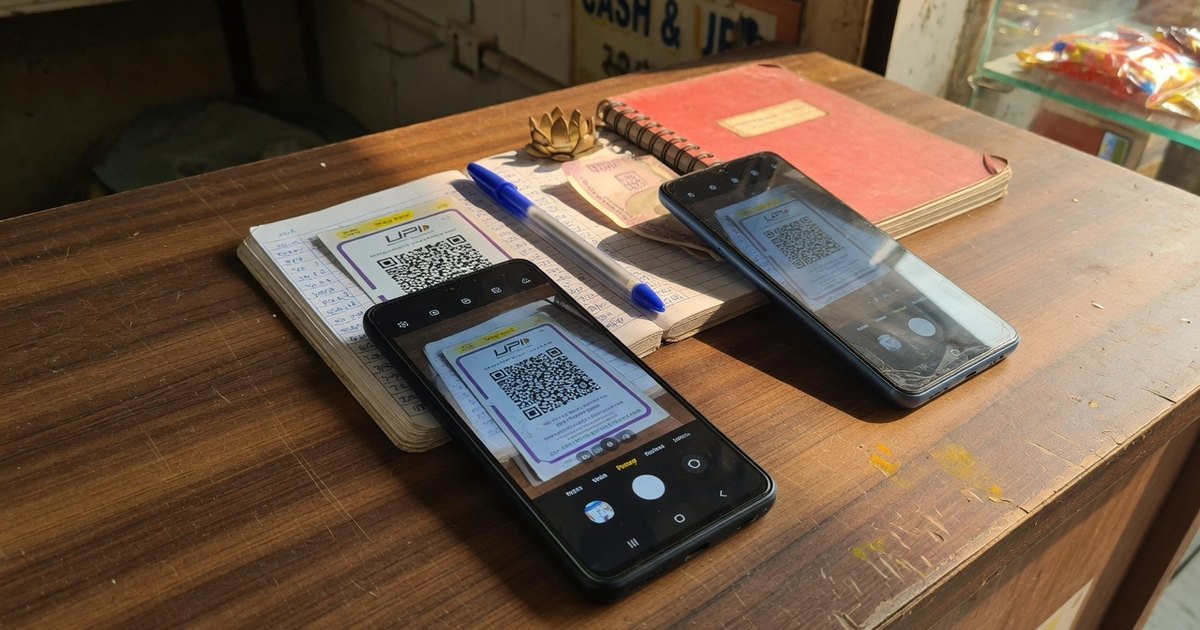 Two phones on a wooden shop desk photographing a printed UPI QR card to read the encoded VPA